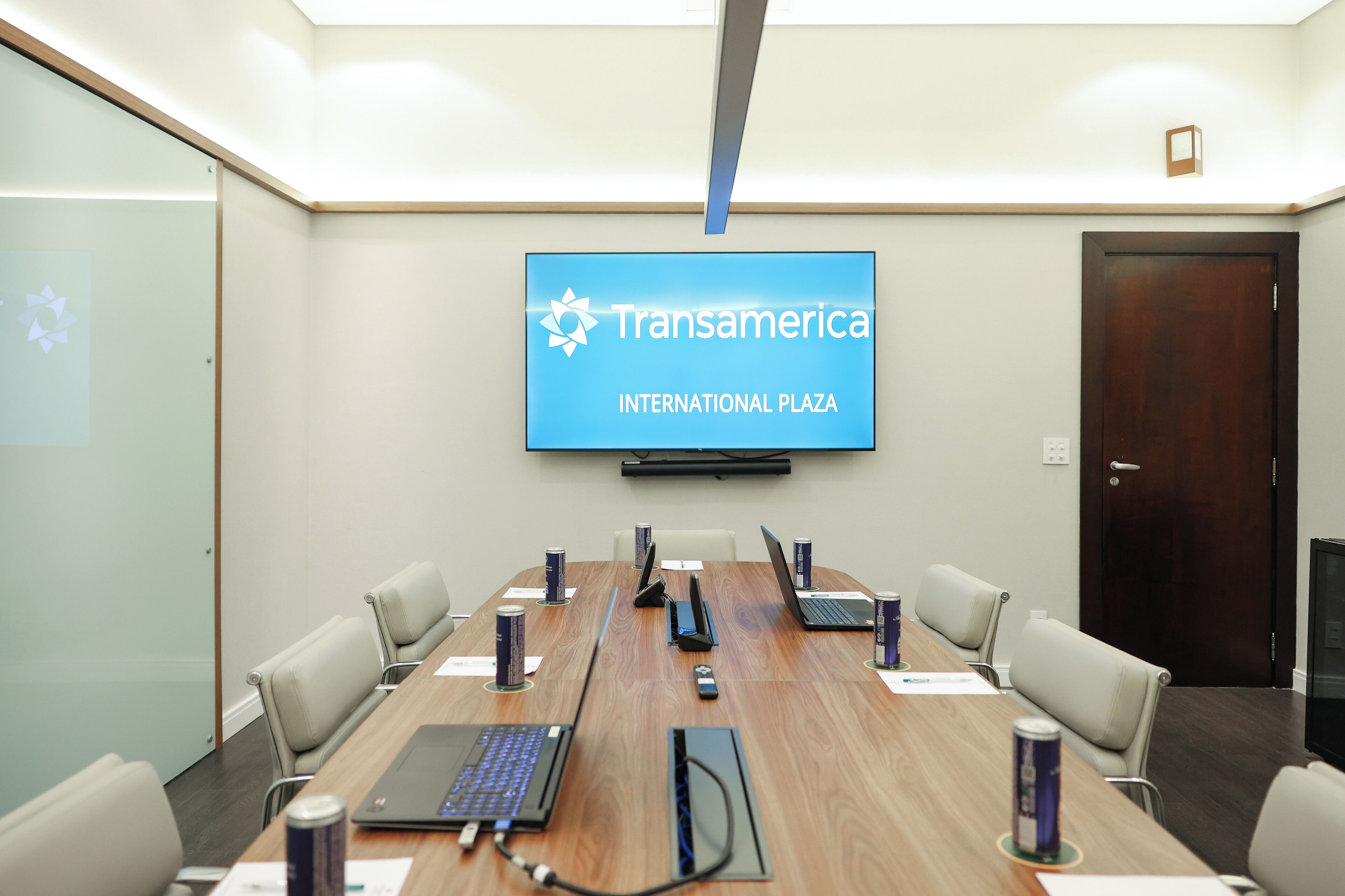 Sala de conferências com uma tela grande exibindo 'Transamerica International Plaza' e uma mesa de madeira com laptops, cadeiras e latas de bebida.