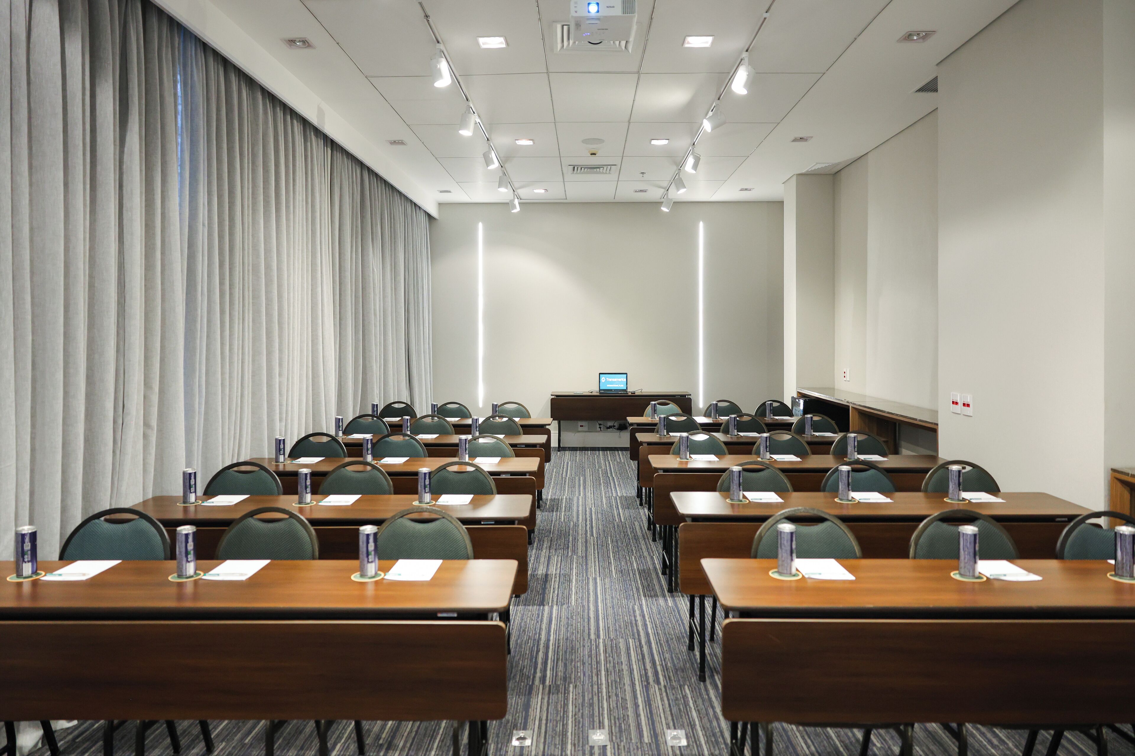 Un salón de conferencias con filas de mesas y sillas, una pantalla de proyector y un panel de control.