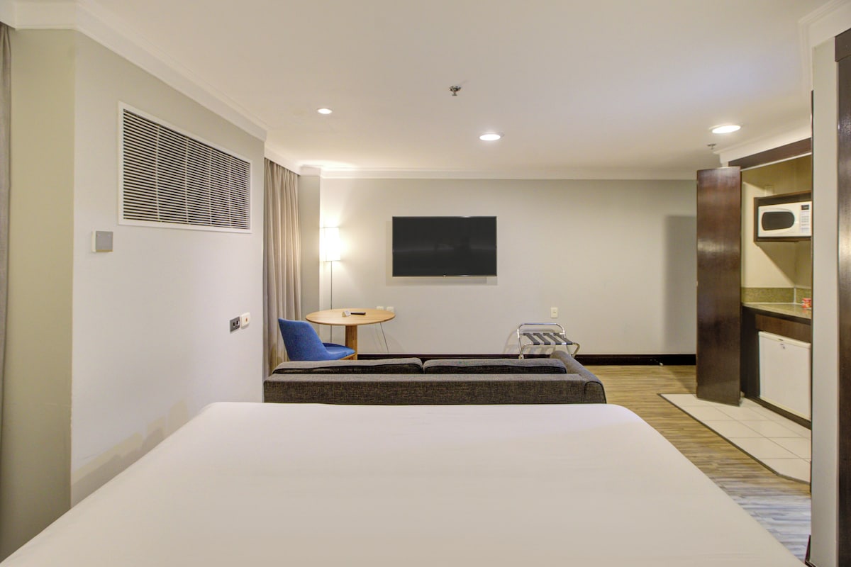 A hotel room with a bed, a small table, a chair, a TV, a microwave, and a kitchenette.