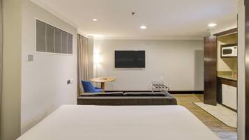 Quarto de hotel com cama, mesa pequena, cadeira, TV, micro-ondas e cozinha compacta.