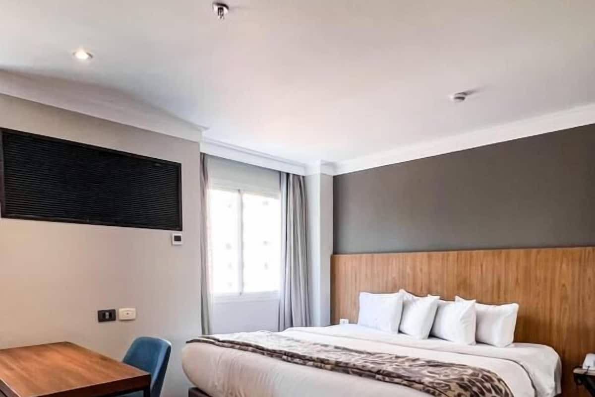 Une chambre d’hôtel moderne avec un plancher en bois, un lit avec du linge de maison blanc, un bureau avec une chaise et une grande fenêtre avec des rideaux.