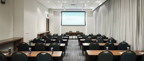 Un salón de conferencias con filas de mesas y sillas, una pantalla de proyector mostrando 'Transamerica International Plaza', y una cortina grande a la derecha.