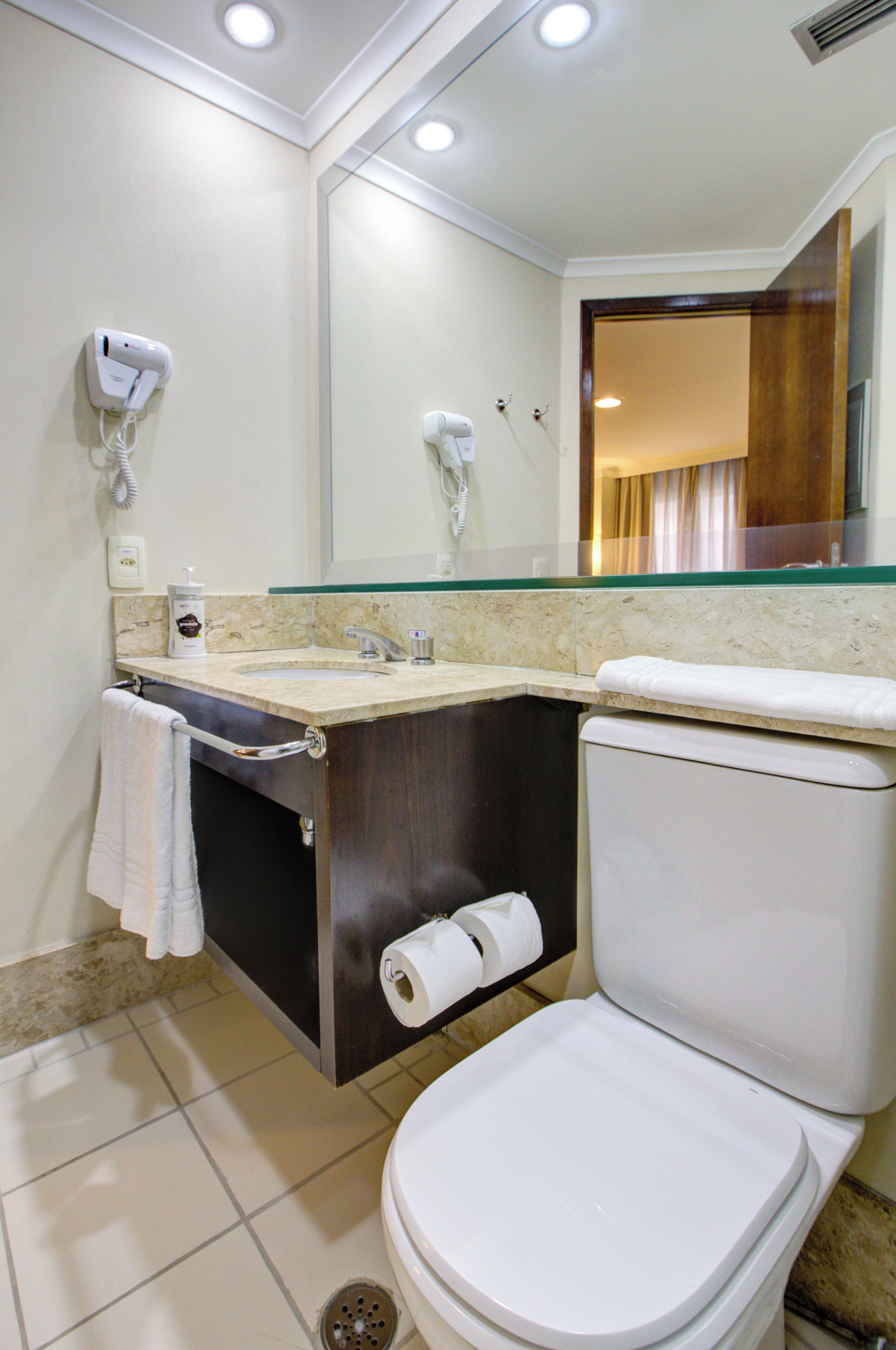 A bathroom with a toilet, sink, and mirror. There are two hair dryers mounted on the wall, a towel hanging on the wall, and a toilet paper roll on the wall.