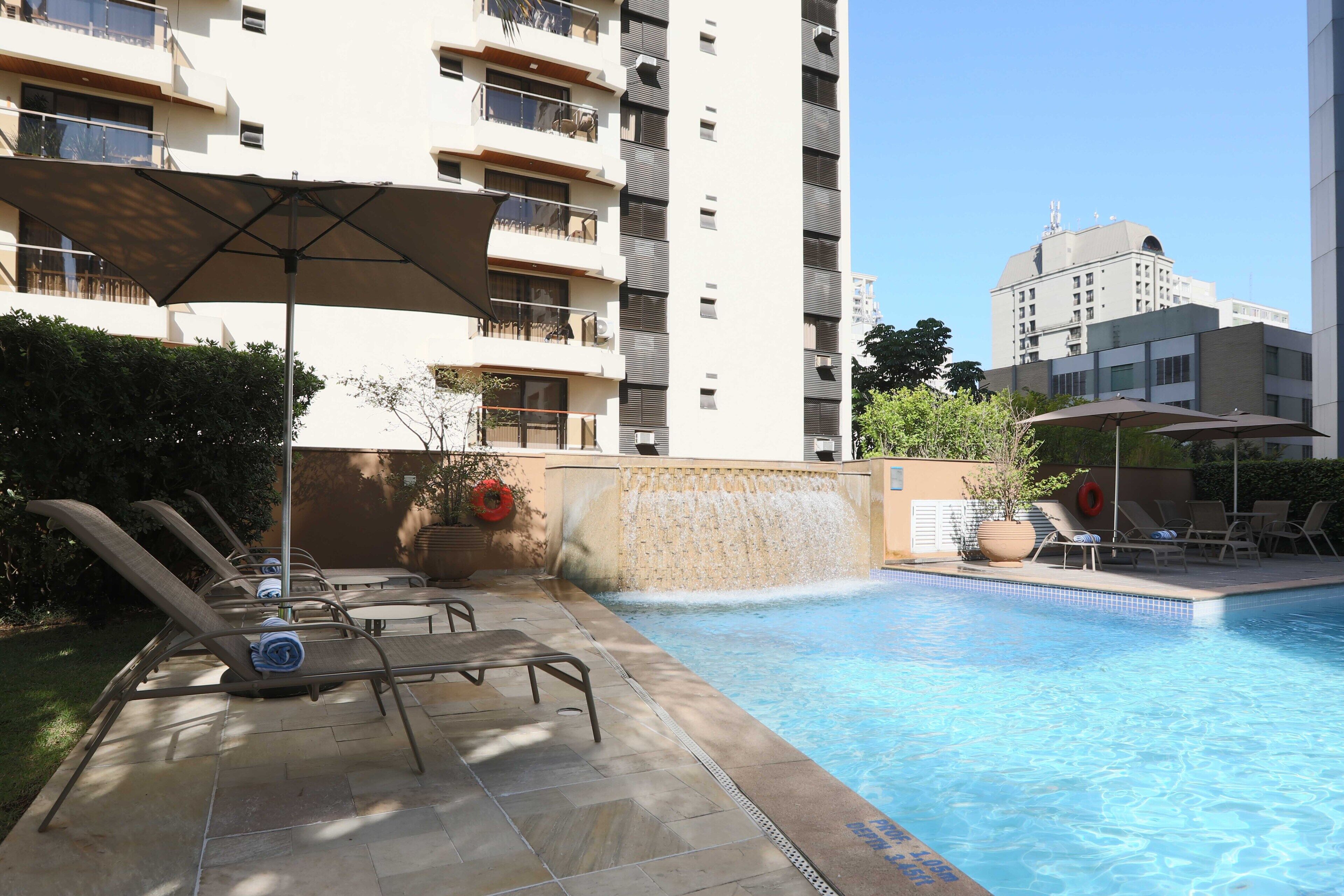 Una piscina con sillones de descanso y sombrillas, rodeada por un edificio alto.