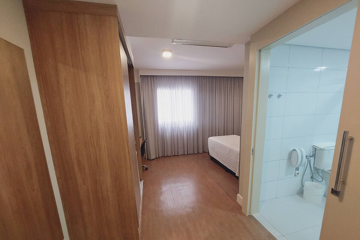 Ett hotellrum med en dörr av trä, en säng, en duschkabin med glasväggar och ett fönster med gardiner.