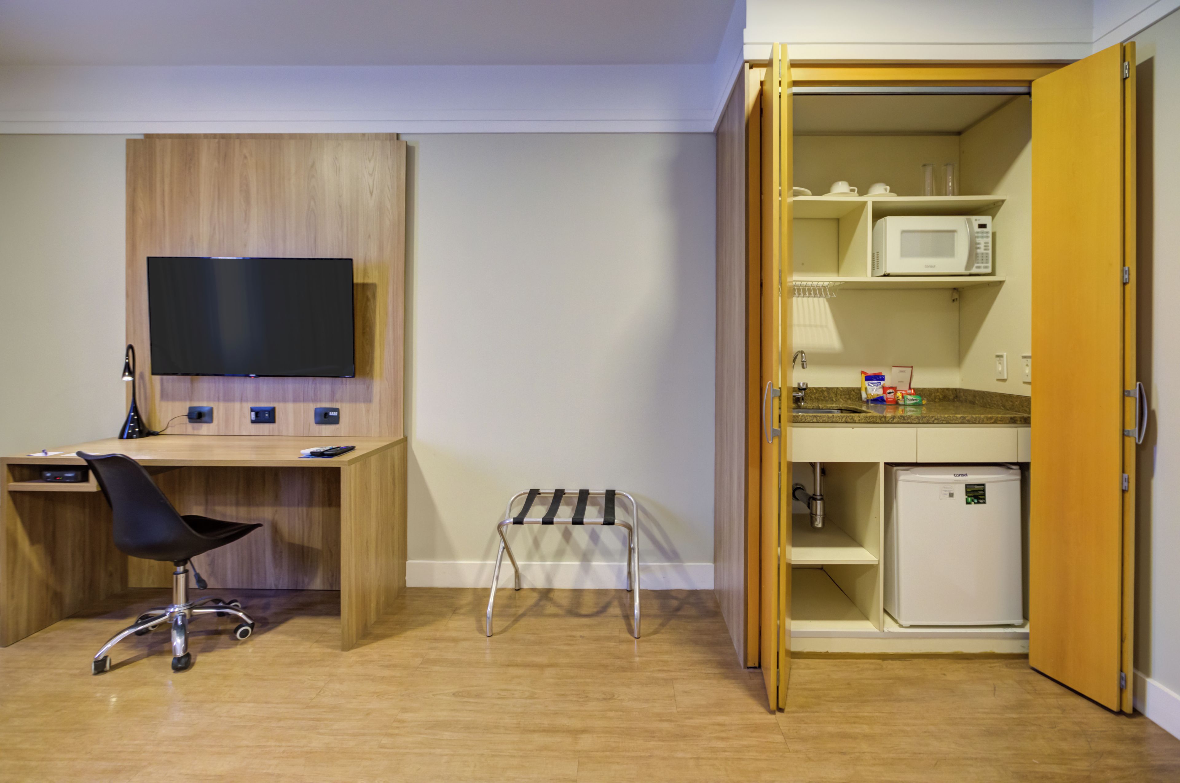 Quarto compacto com escrivaninha de madeira, uma cadeira, televisão, um banquinho pequeno e uma cozinha compacta com micro-ondas e geladeira.
