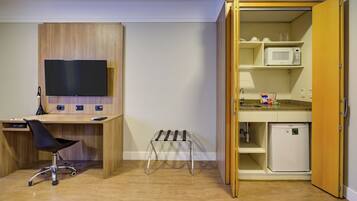 Quarto compacto com escrivaninha de madeira, uma cadeira, televisão, um banquinho pequeno e uma cozinha compacta com micro-ondas e geladeira.