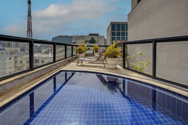 A rooftop pool with clear blue tiles, surrounded by lounge chairs and potted plants, overlooking a cityscape.