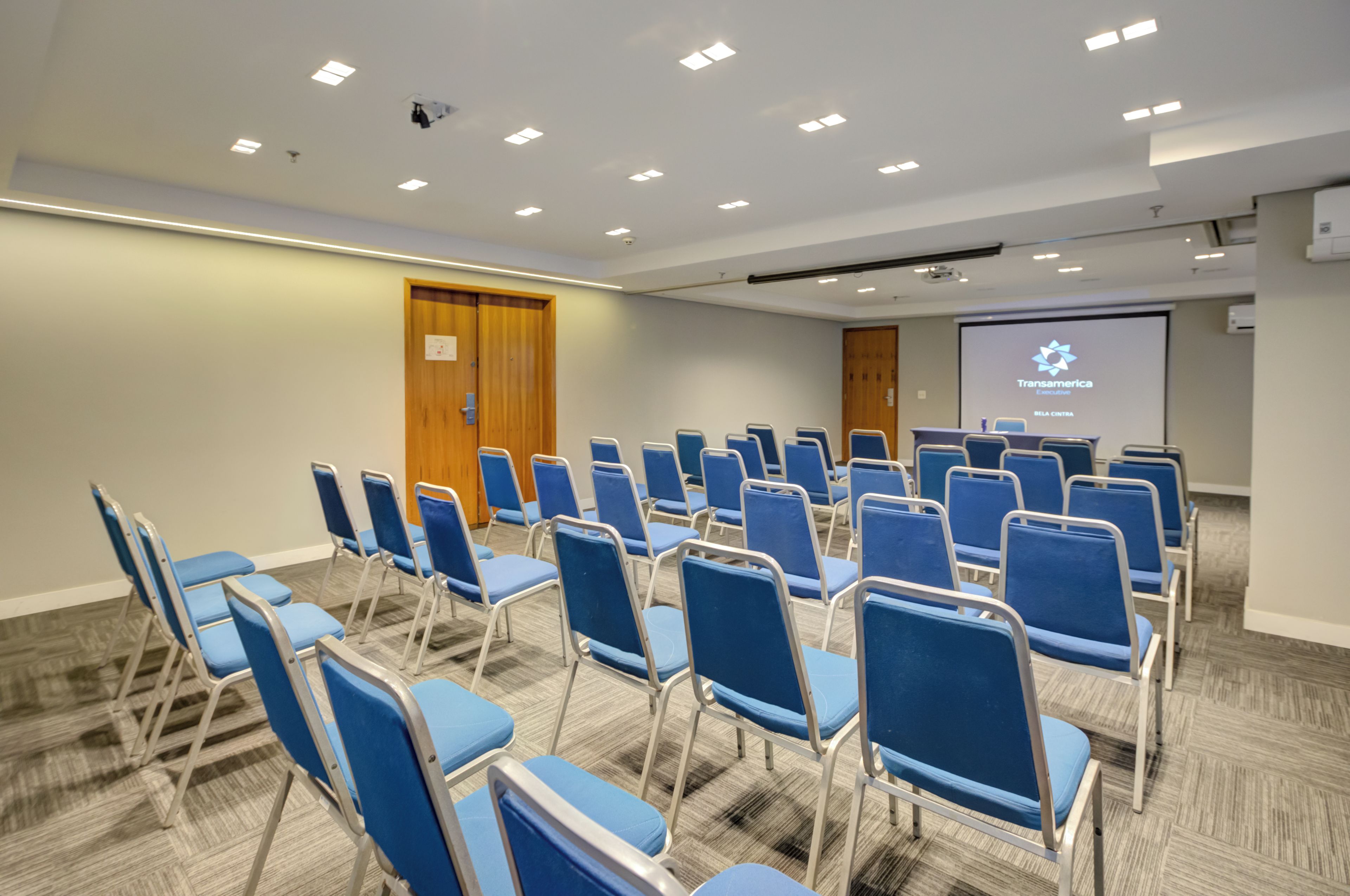 Un salón de conferencias con hileras de sillas azules, una pantalla de proyección y una puerta de madera.