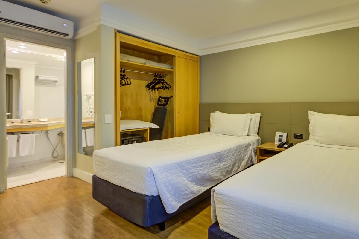 A hotel room with two beds, a wooden wardrobe, a mirror, and a bathroom with a sink and towel rack.