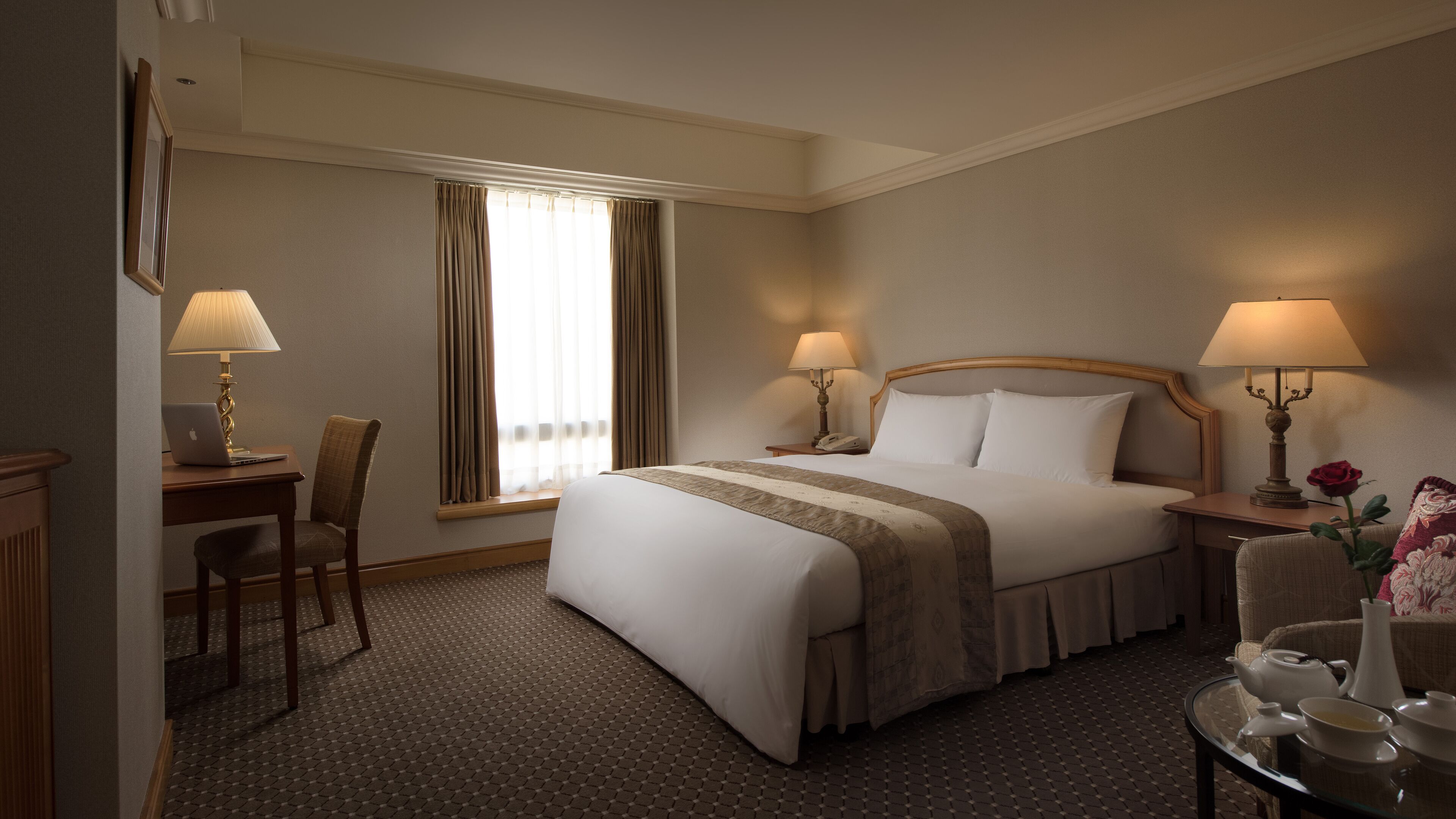 A hotel room with a large bed, two bedside tables with lamps, a desk with a laptop, a chair, and a small table with a vase and teapot.