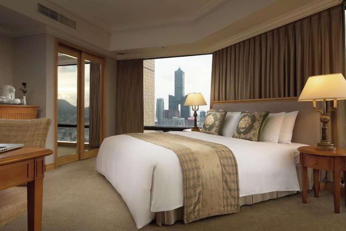 Habitación de hotel con una cama grande, un escritorio, una silla, una lámpara y vistas a un horizonte urbano.