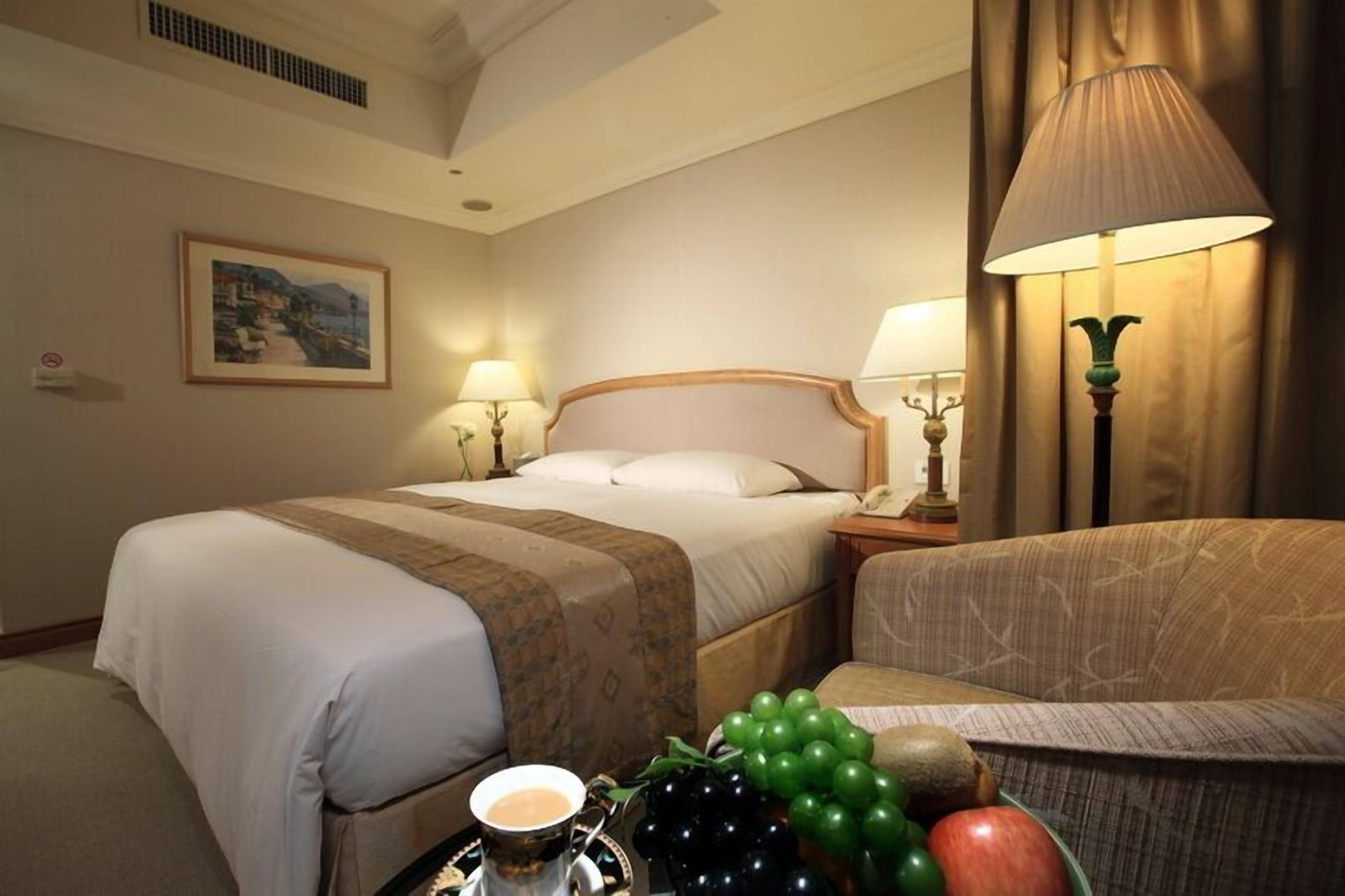 A hotel room with a bed, two bedside lamps, a nightstand, a chair, and a table with fruits and a cup.