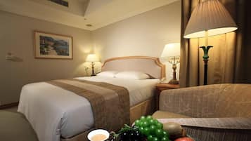 A hotel room with a bed, two bedside lamps, a nightstand, a chair, and a table with fruits and a cup.