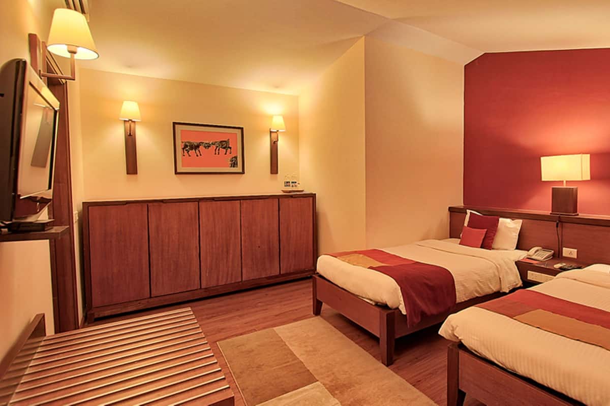 A hotel room with two beds, a wooden headboard, a desk, a lamp, and a framed picture on the wall.