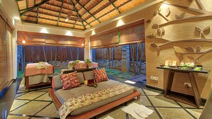 A spacious room with a high ceiling, a bed with a patterned bedspread, a seating area with cushions, a small table with a plant, and a decorative wall with leaf motifs. - Angsana Oasis Spa & Resort (Bengaluru)