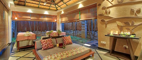 A spacious room with a high ceiling, a bed with a patterned bedspread, a seating area with cushions, a small table with a plant, and a decorative wall with leaf motifs.