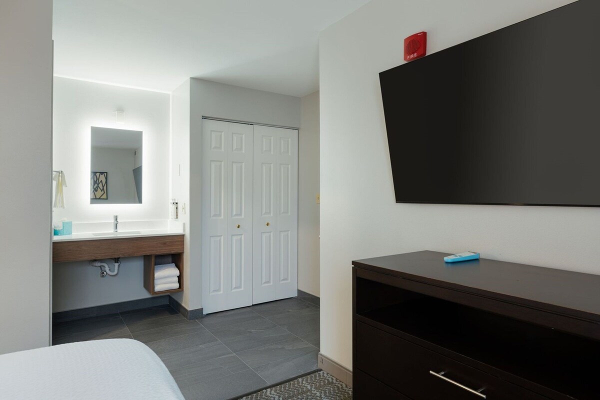 A modern hotel bathroom with a white vanity, a large mirror, a dark wooden dresser, and a flat-screen TV mounted on the wall.