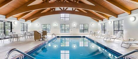 An indoor swimming pool with wooden beams, clear poolside chairs, and a tiled deck.