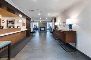 A hotel reception area with a wooden reception desk, a seating area with blue chairs, and a fireplace in the background. - Staybridge Suites Madison East by IHG (Madison)