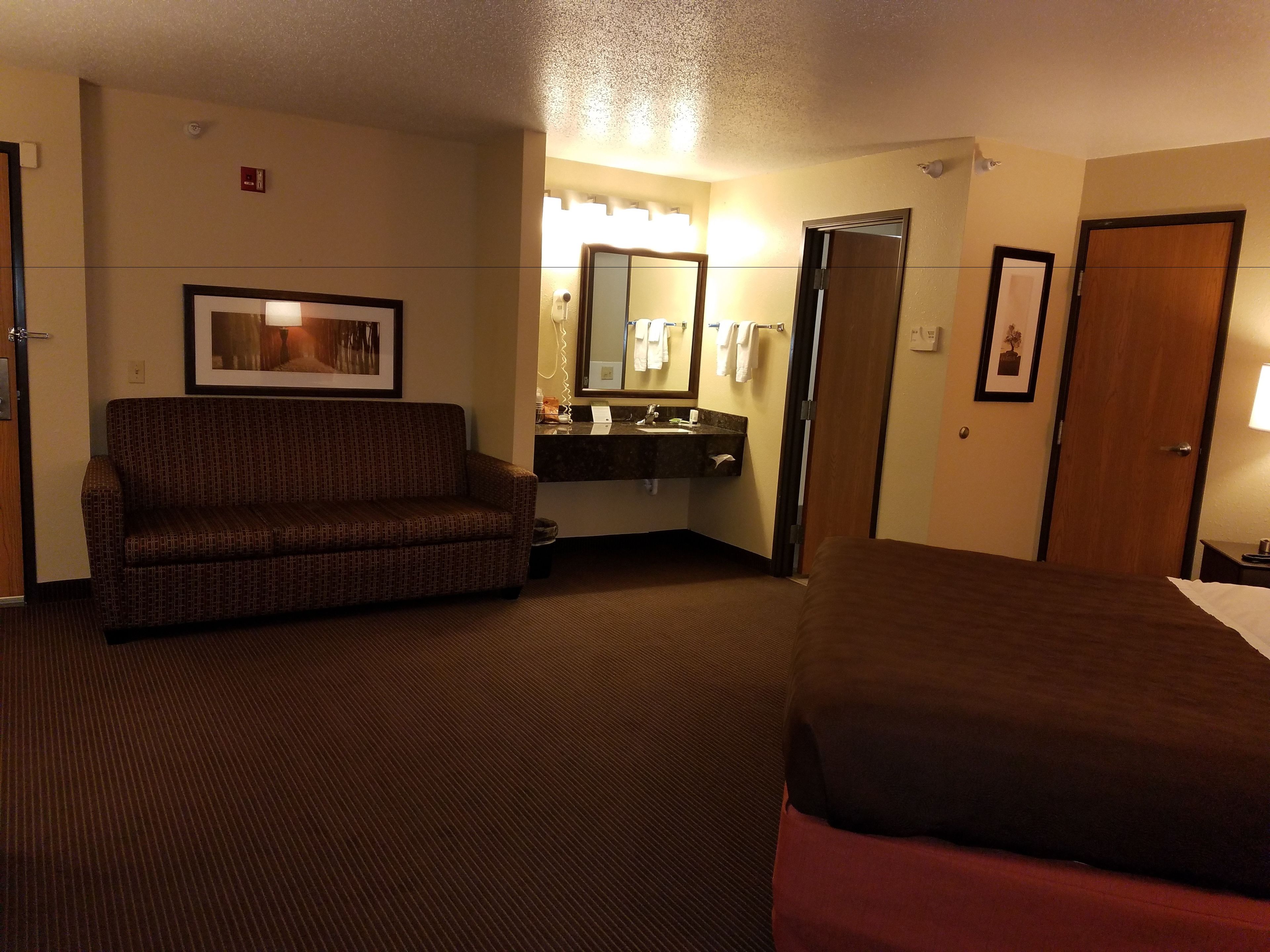A hotel room with a bed, a sofa, a bathroom with a sink and mirror, and a framed picture on the wall.