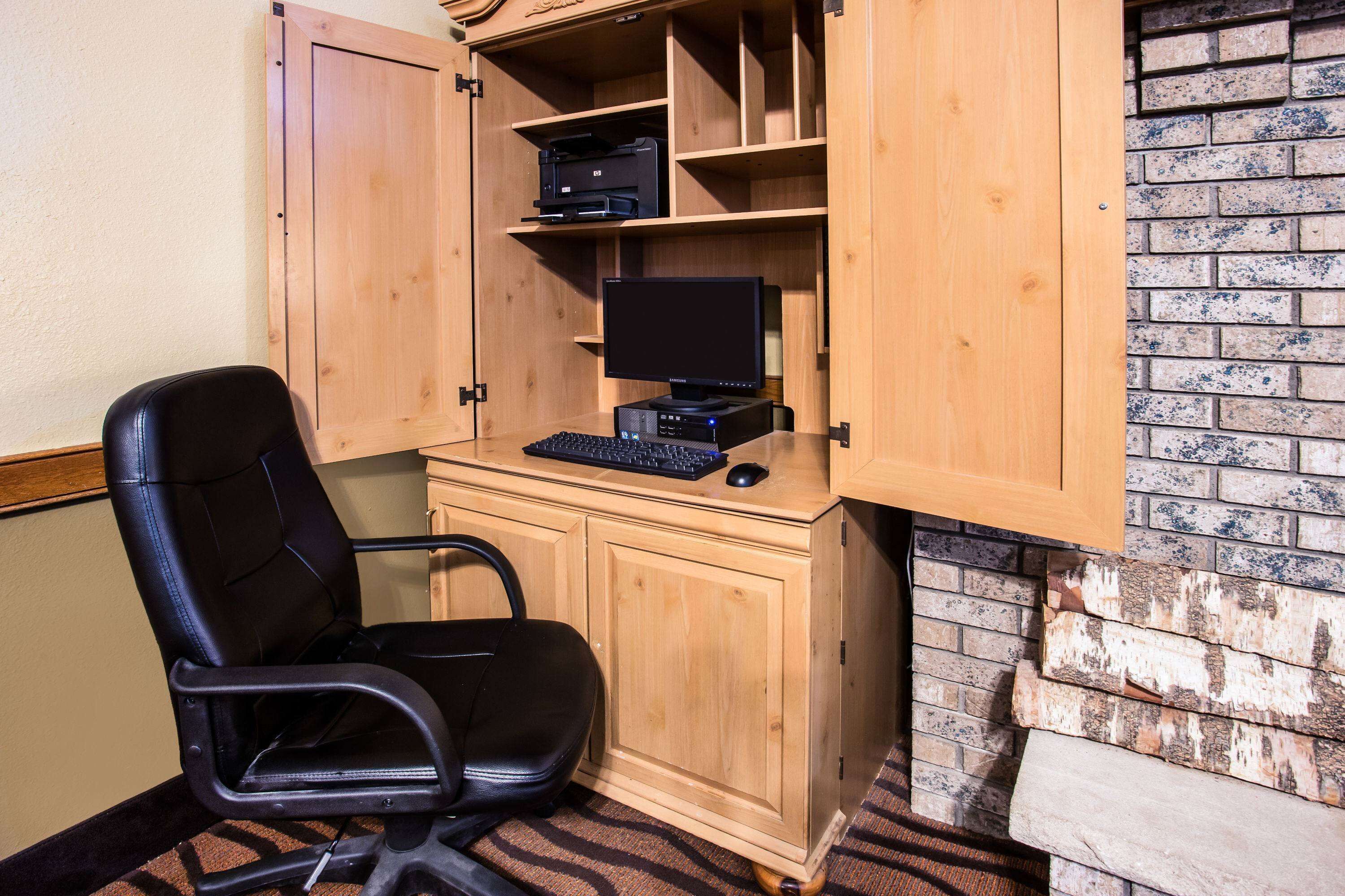 Un escritorio de madera con una computadora, una silla negra y una impresora sobre estanterías.