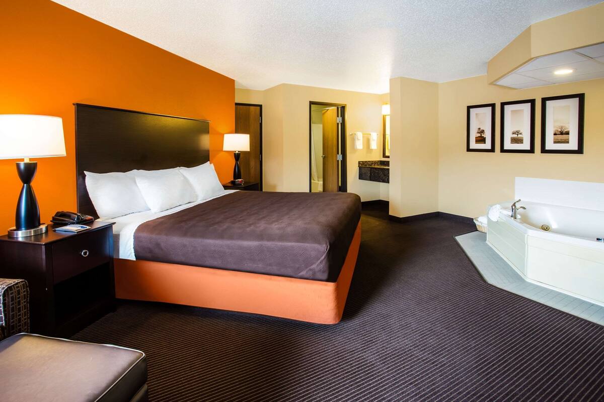 A hotel room with a large bed, a bedside table with a lamp, a bathroom with a bathtub, and framed pictures on the wall.