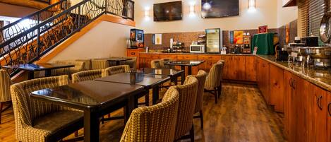 A restaurant with a wooden staircase, wicker chairs, a dining table, a kitchen area with a microwave, and a TV mounted on the wall.