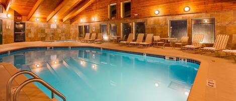 An indoor swimming pool with a wooden ceiling, lounge chairs, and a tiled wall.