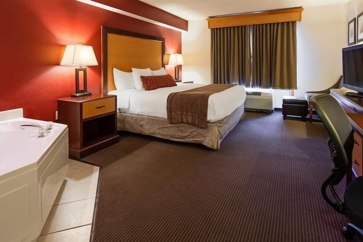 A hotel room with a bed, a desk, a television, a desk lamp, a chair, a desk, a nightstand, a lamp, a window with curtains, and a bathtub.
