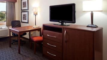 A hotel room with a wooden desk, a television, a lamp, a chair, and a dresser with drawers.