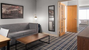 A hotel room with a grey sofa, a wooden coffee table, a framed picture on the wall, a floor lamp, and a wooden door leading to a bathroom. - AmericInn by Wyndham Grand Forks (Grand Forks)