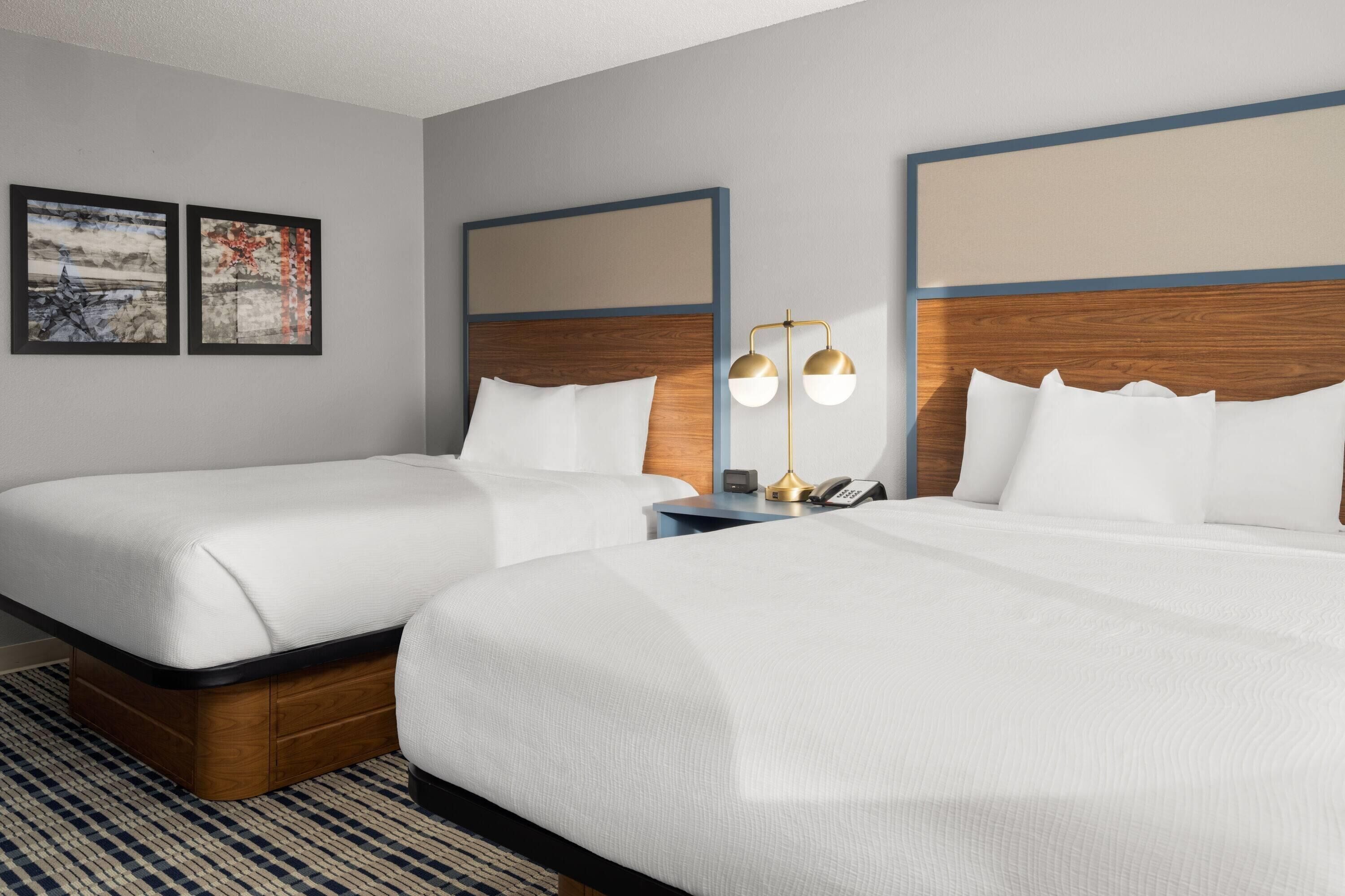 A hotel room with two beds, a nightstand, a lamp, and two framed pictures on the wall.