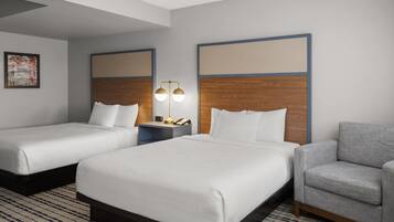 A hotel room with two beds, a gray armchair, a nightstand with a lamp, and a wooden headboard.