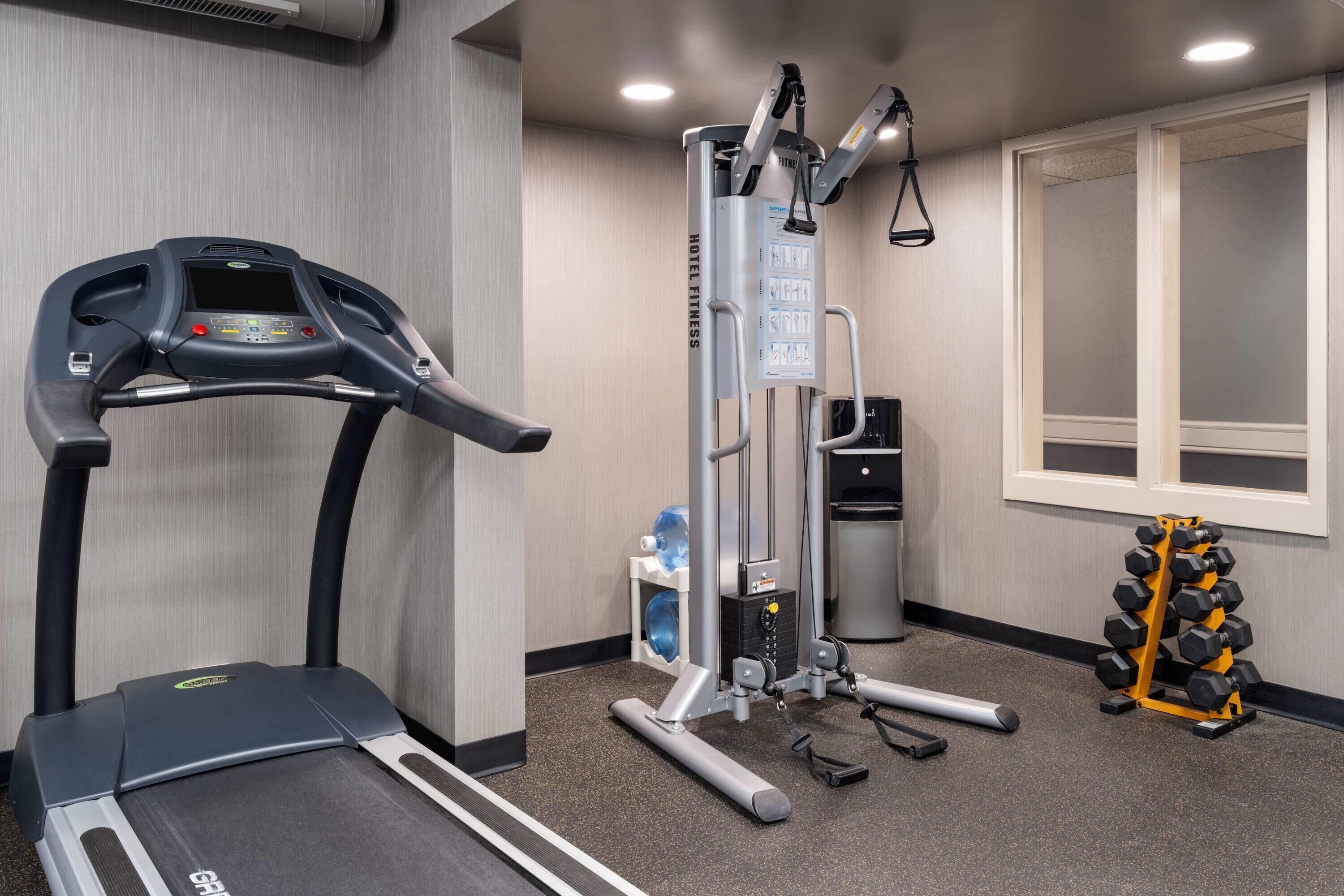 Ein Fitnessraum eines Hotels mit Laufband, Multifunktionsgerät, Kurzhanteln und Wasserspender.