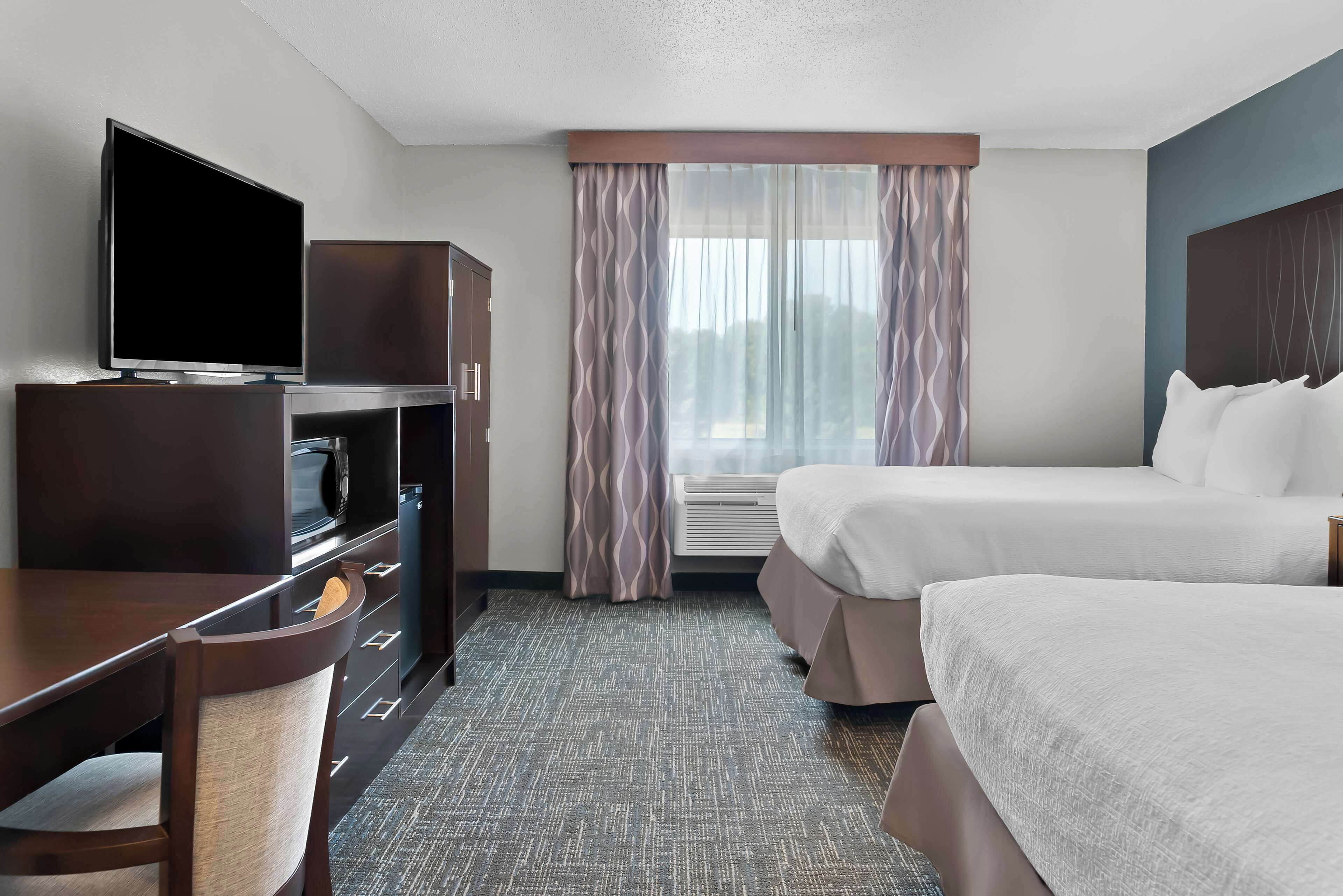 A hotel room with a TV, a desk with a microwave, two beds, and a window with curtains.