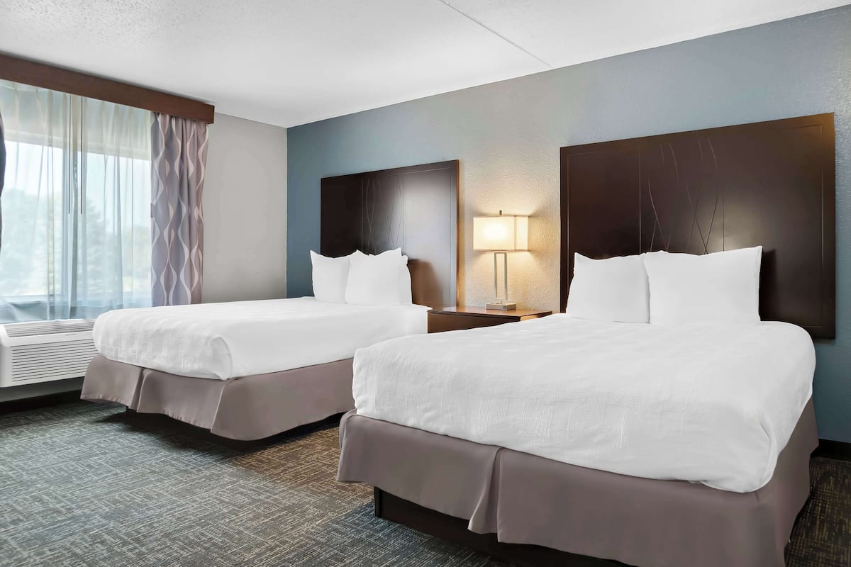 A hotel room with two beds, a nightstand with a lamp, a window with curtains, and a wall-mounted air conditioner.