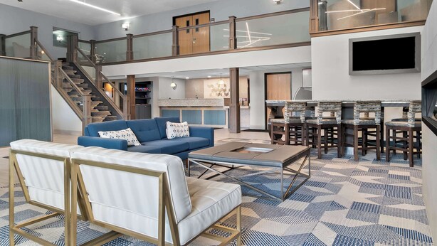Amplia sala de estar con un sofá azul, un sillón blanco y una mesa de centro. Una escalera conduce a un nivel superior con balcón.