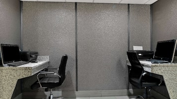 A modern office space with two desks, each with a computer and a printer. The room has a textured gray wall and a tiled floor.