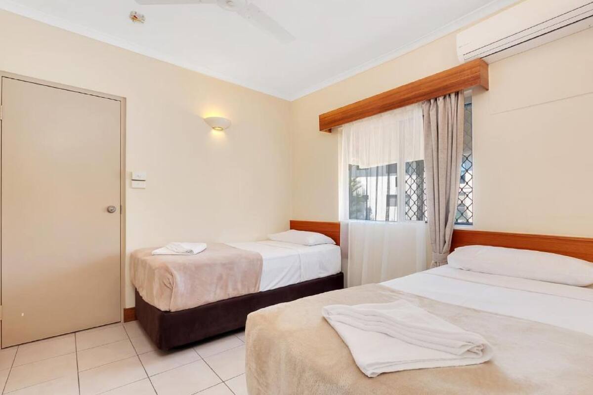 A hotel room with two beds, a wardrobe, a window with curtains, and a wall-mounted light.