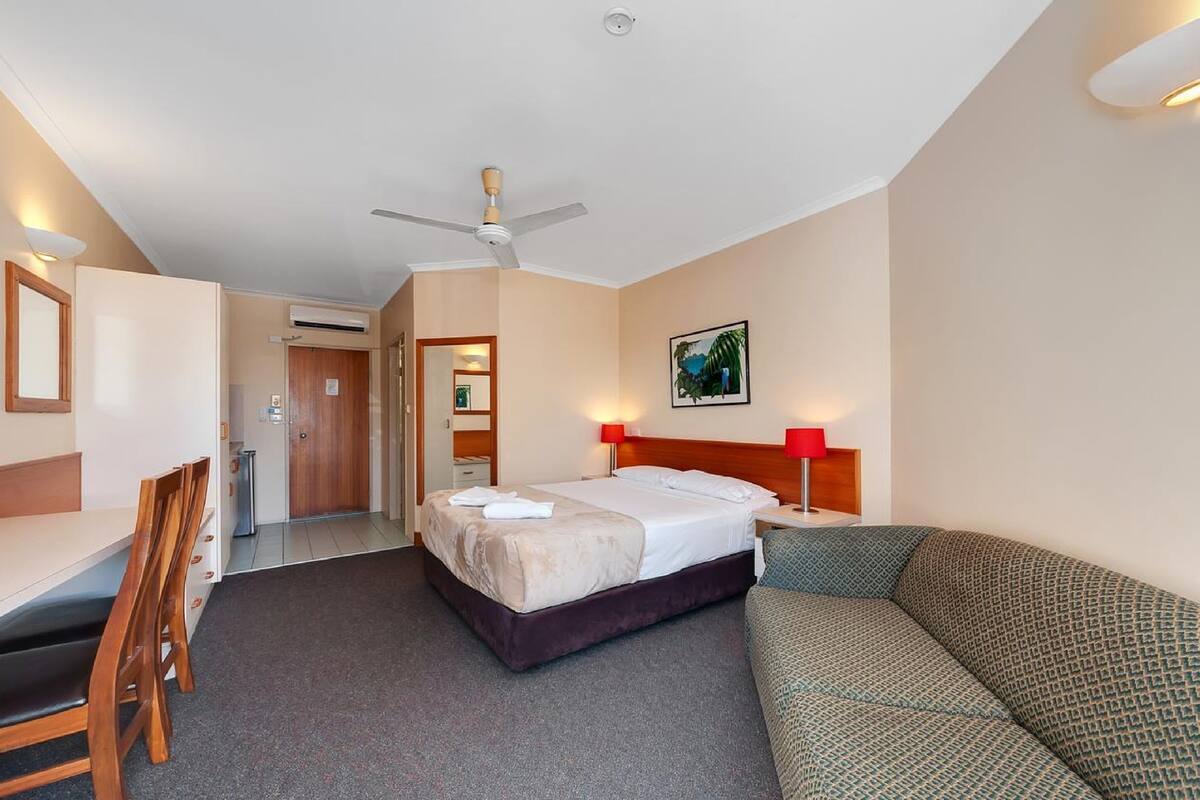 A hotel room with a bed, a desk, a chair, a sofa, a ceiling fan, and a painting on the wall.