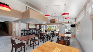A spacious indoor dining area with multiple wooden tables and chairs, red pendant lighting, and large windows allowing natural light.