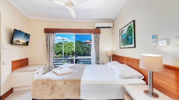 A hotel room with a bed, a nightstand, a desk, a ceiling fan, a window with curtains, and a view of greenery outside.