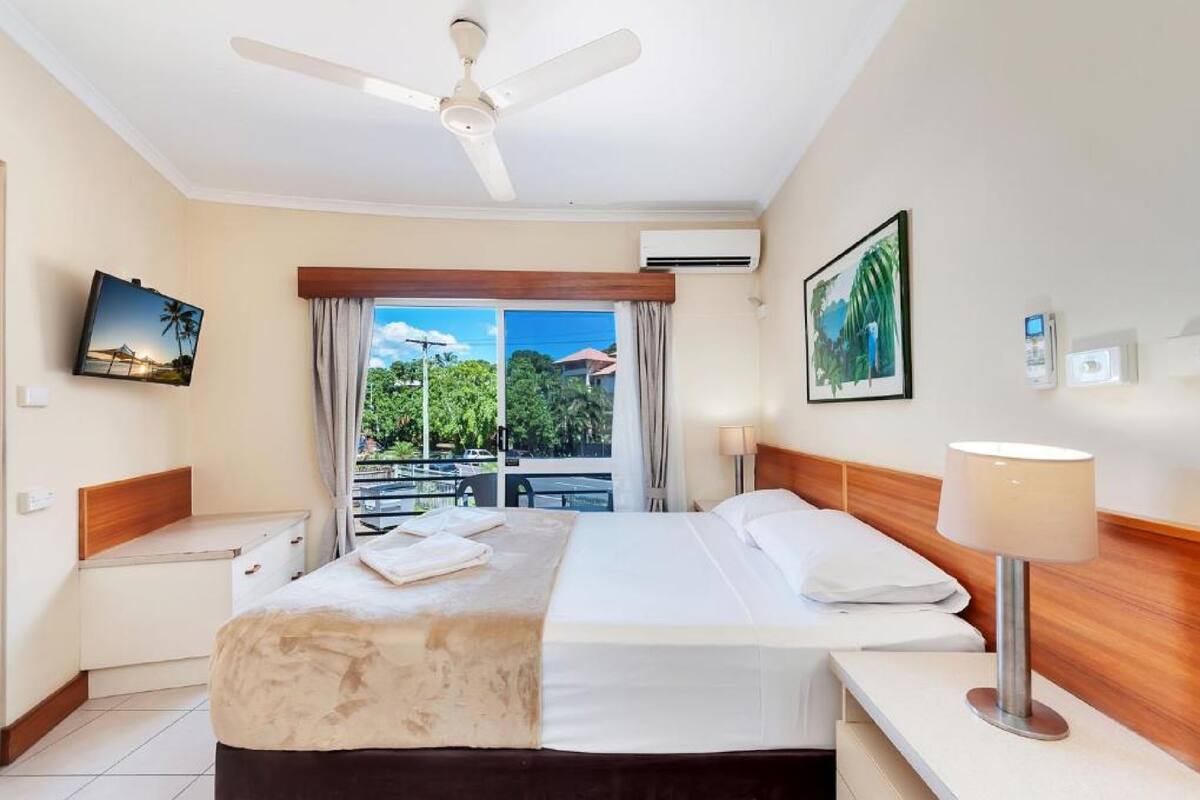 A hotel room with a bed, a nightstand, a desk, a ceiling fan, a window with curtains, and a view of greenery outside.