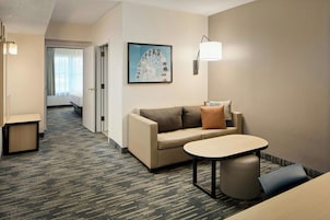 Habitación de hotel con sofá, una mesita, lámpara, un cuadro en la pared y piso alfombrado.