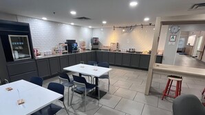 A modern kitchen with white brick walls, a dining area with white tables and blue chairs, and a small bar area with a stool. - City Express by Marriott Louisville Southeast (Louisville)