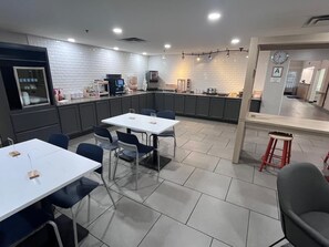 A modern kitchen with white brick walls, a dining area with white tables and blue chairs, and a small bar area with a stool. - City Express by Marriott Louisville Southeast (Louisville)