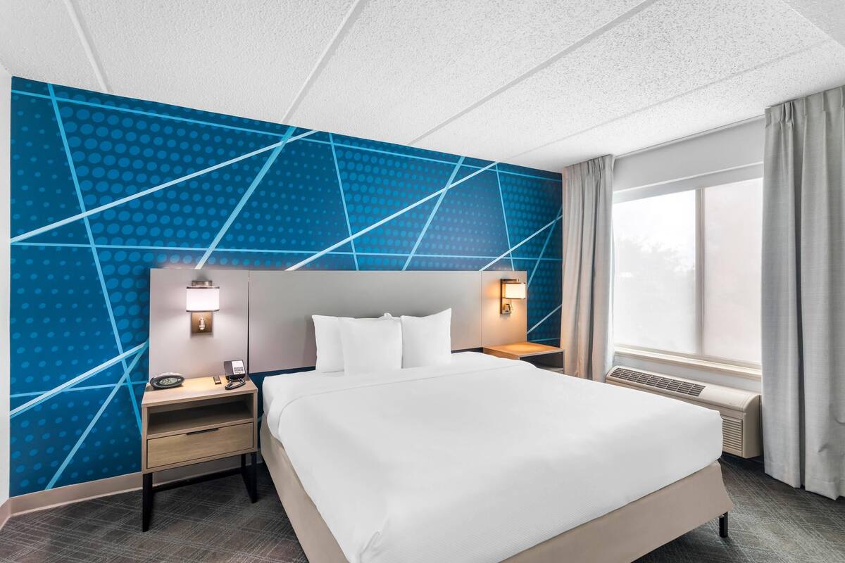A hotel room with a large bed, a nightstand with a lamp, a wall-mounted heater, and a window with curtains.