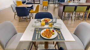 Ein Essbereich mit Tischen und Stühlen, Frühstücksteller mit Waffeln und Obst, sowie ein an der Wand befestigter Fernseher.