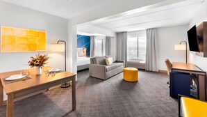 Ein Hotelzimmer mit Essbereich, einer Couch, einem Fernseher und einem Fenster mit Vorhängen.